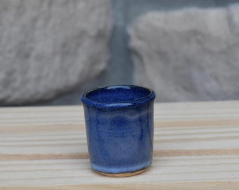 May include: A small, cylindrical ceramic cup featuring a deep cobalt blue glaze that transitions to a lighter, sky blue hue near the base. The rim is smooth and rounded, while the exterior surface displays a glossy, reflective finish throughout.