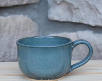 May include: A wide, low-profile ceramic mug with a smooth, matte blue-grey glaze. The mug features a rounded body, a sturdy curved handle on the right side, and a visible unglazed tan base that contrasts with the blue-grey finish of the vessel.