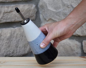 May include: A ceramic oil cruet featuring a tapered, conical shape with a three-tone color scheme. The top section is white, the middle is a speckled light blue, and the base is solid black. It includes a stainless steel pour spout with a black cap.
