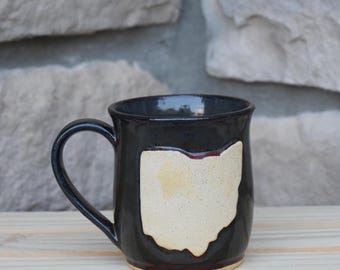 May include: A dark navy blue ceramic mug with a glossy finish featuring a prominent, light cream-colored cutout in the shape of the state of Ohio on the front. The mug has a curved handle and a smooth, rounded rim, showcasing a handcrafted design.