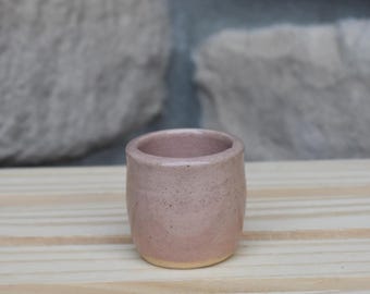 May include: A small, cylindrical ceramic cup with a speckled, dusty pink glaze. The vessel features a smooth, rounded rim and a slightly tapered base that reveals a natural, unglazed tan clay finish at the very bottom edge of the exterior.