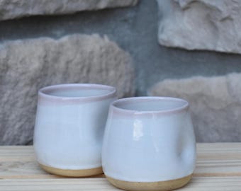 May include: Two small, rounded ceramic cups with a smooth white glaze and a subtle pinkish-tan rim. The base of each cup is unglazed, revealing a natural tan clay color. The cups have a simple, handleless design with a slightly indented side.