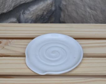 May include: A round, white ceramic soap dish featuring a raised spiral pattern embossed into the center. The dish has a smooth, glossy finish and a small indentation along the edge, designed to hold a bar of soap securely on a flat surface.