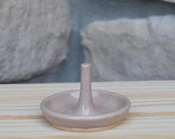 May include: A light pink ceramic ring holder featuring a circular base and a central vertical post. The surface of the pottery has a subtle speckled texture with a glossy finish, and the base shows a natural, unglazed tan clay rim at the bottom.