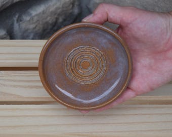 May include: A small, round ceramic grater plate featuring a glazed finish in shades of brown and muted blue. The center of the plate displays a raised, circular textured pattern designed for grating, surrounded by a smooth, slightly elevated rim.