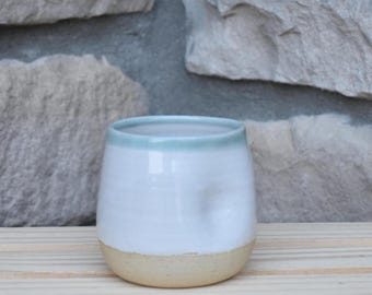 May include: A small, rounded ceramic cup featuring a smooth white glaze on the upper portion and a natural, unglazed beige clay base. The rim is finished with a thin, soft mint green band, and the body has a subtle indentation on one side.