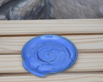 May include: A round, blue ceramic spoon rest featuring a prominent spiral design etched into the center. The glaze has a glossy finish with subtle variations in color intensity and a slightly raised edge to contain liquids or food residue.