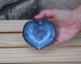 May include: A small, heart-shaped ceramic dish featuring a speckled, deep blue glaze with a smooth, glossy finish. The bowl has a slightly indented top edge and a curved, concave center, showcasing a handcrafted appearance with subtle color variations.