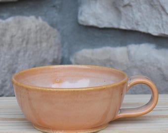 May include: A wide, shallow ceramic mug with a smooth, glossy peach-colored glaze. The mug features a rounded body, a short, sturdy handle on the right side, and a slightly darker, speckled rim. The surface has subtle variations in color tone.
