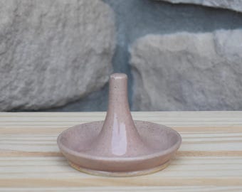 May include: A dusty rose ceramic ring holder featuring a central conical post rising from a shallow, circular base. The glazed surface has a subtle speckled texture throughout, with a smooth finish and a slightly flared rim on the base.