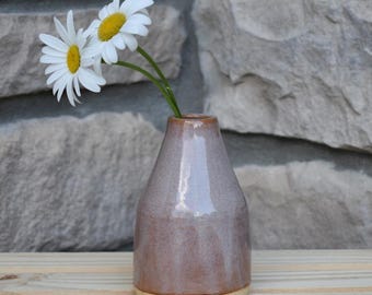 May include: A small, tapered ceramic vase with a glossy, muted mauve-brown glaze and a smooth, unglazed beige base. Two white daisies with bright yellow centers are placed inside the vase, with their green stems extending from the narrow opening.