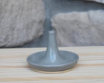 May include: A gray ceramic ring holder featuring a circular base and a central conical post. The piece has a smooth, glossy finish on the top and sides, with a visible unglazed light beige clay rim along the bottom edge of the circular base.