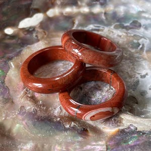 Könnte beinhalten: Drei rote Jaspis-Ringe mit natürlichen Mustern. Die Ringe sind glatt und poliert und weisen Farbvariationen von tiefem Rot bis zu helleren Farbtönen mit weißen und braunen Einschlüssen auf. Die Ringe werden auf einer perlmuttartigen Oberfläche präsentiert.