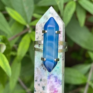 May include: A silver ring featuring a striking blue crystal. The crystal is a long, pointed shape, and the ring has a simple, elegant design. The crystal is mounted on a multi-coloured stone obelisk, with green foliage in the background.