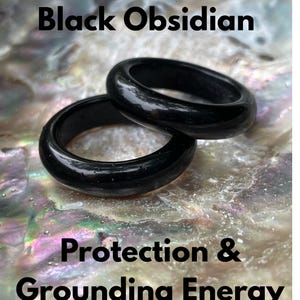 Könnte beinhalten: Zwei polierte schwarze Obsidianringe liegen auf einer perlmuttartigen Oberfläche. Die Ringe sind glatt und glänzend und reflektieren das Licht. Der Text auf dem Bild lautet "Black Obsidian" und "Protection & Grounding Energy."