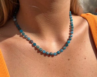 Puede incluir: Un collar de cuentas turquesas con pequeñas cuentas facetadas. El collar está ensartado en una cadena dorada y descansa sobre el pecho de una persona. El collar es de estilo gargantilla. Las cuentas miden aproximadamente 0,5 cm de diámetro.