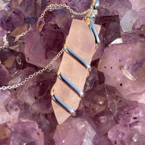 Op de afbeelding: Een roze rozenkwarts hanger gewikkeld in zilverdraad, hangend aan een zilveren ketting. De hanger ligt op een bed van amethist kristallen.