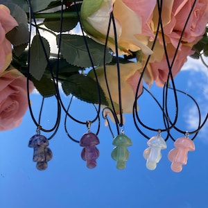 Puede incluir: Una colección de colgantes de cristal con forma de hongo sobre cordones negros. Los colgantes son de varios colores, incluyendo azul, morado, verde, blanco y rosa. El fondo es una superficie reflectante con un reflejo del cielo y flores.
