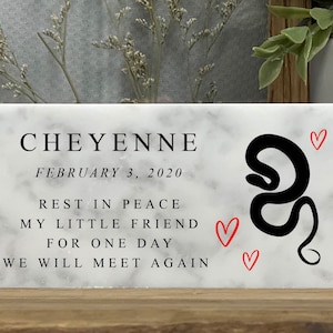 May include: White marble memorial plaque with black text that reads "CHEYENNE, FEBRUARY 3, 2020, REST IN PEACE, MY LITTLE FRIEND FOR ONE DAY, WE WILL MEET AGAIN". A black snake silhouette is on the right side of the plaque with three red hearts.