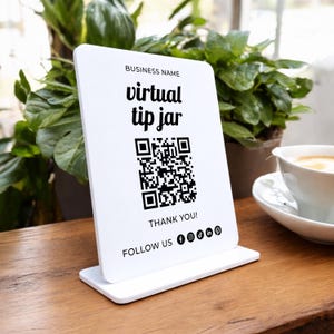 Peut inclure: Panneau blanc rectangulaire avec l'inscription "virtual tip jar" et un code QR noir. Le panneau affiche également le texte "THANK YOU!" et des icônes de réseaux sociaux. Le panneau est posé sur une surface en bois.