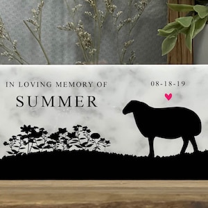 May include: A white marble memorial plaque with black text that reads "In loving memory of Summer 08-18-19". A black silhouette of a sheep with a pink heart above it is in the upper right corner. A row of black silhouette flowers are at the bottom of the plaque.