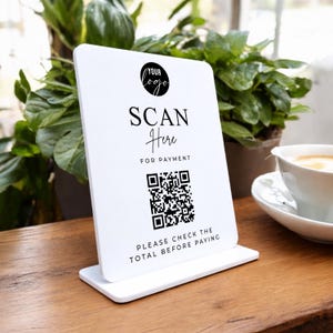 May include: A white QR code payment sign with the text "SCAN Here FOR PAYMENT" and a place for a logo. The sign is on a wooden table with a cup of coffee and plants in the background. The sign also says "PLEASE CHECK THE TOTAL BEFORE PAYING."