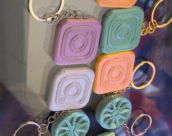 May include: A collection of colorful keychains featuring various designs. The keychains include Instagram logo squares in shades of orange, green, purple, and white, along with wheel and cassette tape designs. Each keychain has a metal ring.
