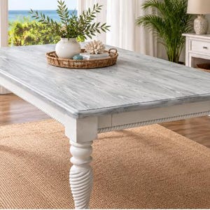 Puede incluir: Una mesa de comedor rectangular con una superficie de madera gris desgastada y una base blanca con cuatro patas gruesas, torneadas y en espiral. La mesa sostiene una bandeja redonda de mimbre trenzado con un jarrón de cerámica blanca.