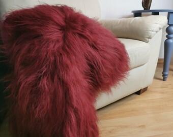 May include: A deep red, shaggy throw blanket draped over a cream-colored armchair. The blanket's texture is long and fluffy, creating a cozy and inviting look. A small, dark mug sits on a blue side table in the background, adding a touch of contrast.