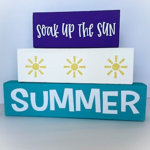 May include: Three wooden blocks stacked on top of each other. The top block is purple and says "Soak Up The Sun" in white letters. The middle block is white with three gold glitter sunbursts. The bottom block is teal blue and says "SUMMER" in white letters.