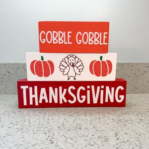 May include: Three stacked wooden blocks with Thanksgiving-themed designs. The top block is orange with "GOBBLE GOBBLE" in white. The middle block is white with red pumpkins and a turkey. The bottom block is red with "THANKSGIVING" in white.