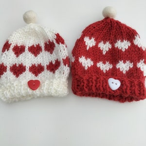 Op de afbeelding: Twee gebreide mutsen met hartjespatronen. De ene muts is wit met rode hartjes en de andere is rood met witte hartjes. Beide mutsen hebben een witte pompon bovenop en een hartvormige knoop.