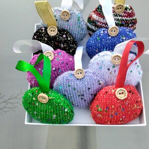 May include: Eight handmade knitted heart decorations with sequins and wooden buttons that say "Handmade with love". The decorations are in various colours, including red, green, blue, purple, pink, black, and white.