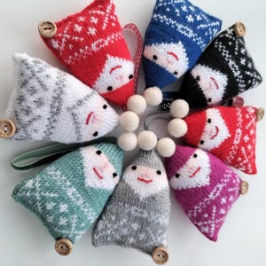 May include: Eight knitted gnome ornaments in various colours and patterns, including red, blue, green, grey, and pink. Each gnome has a white pom-pom hat and a button nose. The gnomes are arranged in a circle around a string of white pom-poms.
