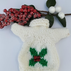 May include: White knitted sweater with a green holly design and red berries. The sweater is a Christmas themed garment.