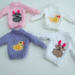 Puede incluir: Cuatro jerséis de punto para muñecas, cada uno con una aplicación de animal diferente. Un jersey es rosa con un gato gris, uno es morado con un pato amarillo, uno es blanco con un pato amarillo y uno es blanco con un conejito marrón.