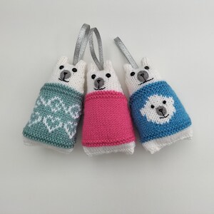 May include: Three white knitted polar bear ornaments with different colored sweaters. One has a teal sweater with a snowflake pattern, one has a pink sweater, and one has a blue sweater with a white polar bear on it.