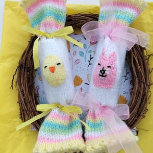 May include: Two knitted Easter egg-shaped gift bags with colourful stripes. One bag features a yellow chick face and the other a pink cat face. Both bags are tied with ribbon.