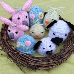 Puede incluir: Una colección de decoraciones de Pascua de punto, incluyendo conejos, pollitos, corderos y huevos, dispuestos en un nido tejido. Las decoraciones están hechas en colores pastel, incluyendo rosa, azul, amarillo y blanco.