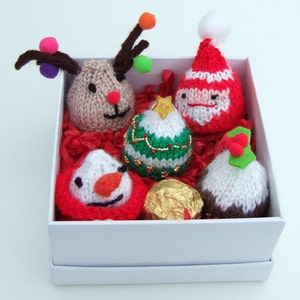 May include: A white gift box filled with six knitted Christmas decorations. The decorations include a reindeer, Santa Claus, a snowman, a Christmas tree, a pudding, and a chocolate ball.