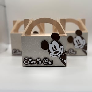 Peut inclure: Trois boîtes à cadeaux gris clair avec des poignées en bois. Chaque boîte présente un motif Mickey Mouse et le texte "Elias is One". Les boîtes ont un motif Mickey Mouse gris clair.