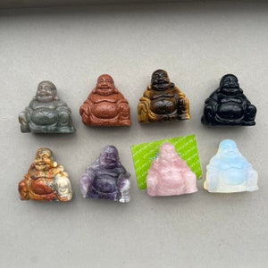 Puede incluir: Siete pequeñas figuritas de Buda talladas en piedra, cada una en un color y material diferente. Las figuritas están dispuestas en fila sobre una superficie gris claro. Los colores incluyen verde, marrón, dorado, negro, morado, rosa y blanco.