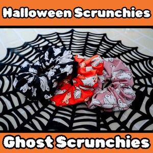 Op de afbeelding: Drie stoffen scrunchies met een spookjespatroon. De scrunchies zijn zwart, rood en roze. Ze liggen op een zwarte spinnenweb-achtergrond.
