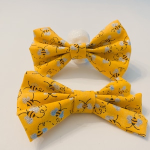 May include: Two yellow fabric bow ties with a bee pattern. The smaller bow tie is in front of the larger bow tie.