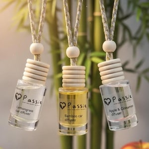 Könnte beinhalten: Drei hängende Auto-Diffusoren mit Holzakzenten und transparenten Glasflaschen. Die Flaschen enthalten Flüssigkeit in verschiedenen Farben und sind mit dem Markennamen "Passie" und Duftbeschreibungen versehen.