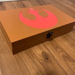 Pu&ograve; includere: Una scatola di legno marrone con un simbolo rosso dell'Alleanza Ribelle sul coperchio. La scatola rettangolare ha una chiusura nera. La scatola &egrave; su una superficie di legno.