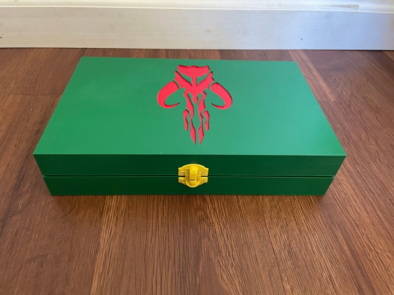 Pu&ograve; includere: Una scatola rettangolare di legno verde con un emblema rosso sul coperchio. La scatola ha una chiusura color oro. L'emblema &egrave; un design stilizzato con linee curve e punte.