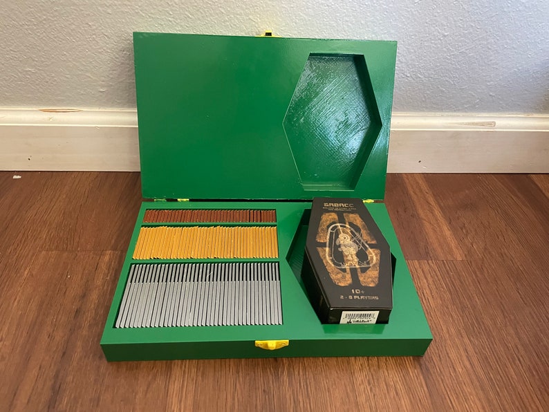 Pu&ograve; includere: Una scatola di legno verde con chiusura dorata, aperta per rivelare carte da gioco di vari colori e una scatola di carte a forma di bara. La scatola a bara ha la parola "SRENCC".