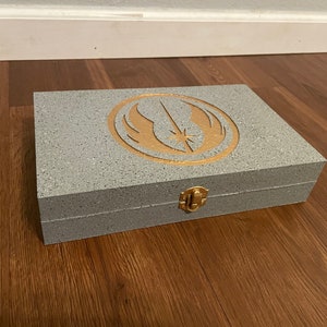 Pu&ograve; includere: Una scatola rettangolare grigia con un emblema dorato sul coperchio. L'emblema presenta un design stilizzato di uccello e stella. La scatola ha una chiusura dorata ed &egrave; posta su una superficie di legno.