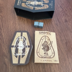 Pu&ograve; includere: Un set di carte Sabacc con una scatola nera a forma di bara, carte, dadi e istruzioni. La scatola e le carte presentano un design grafico oro e bianco. Il gioco &egrave; per 2-8 giocatori, dai 10 anni in su.